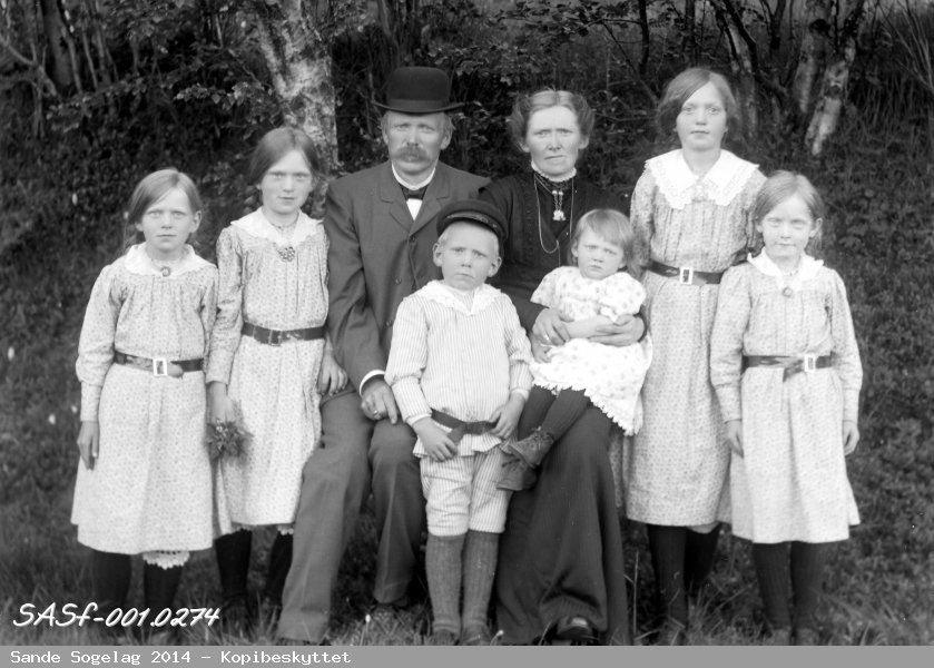 Gammalt bilde av ein familie, mor og far sit med to av barna i fanget og dei 4 andre barna rundt seg.Bildet er tatt ute med bjørkeskog og gras i bakgrunnen.