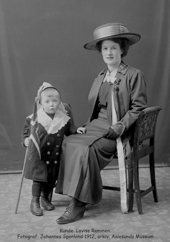 Festkledd kvinne med lang 17. maisløyfe og stor hatt sittande i stol i fotostudio. Ein liten festkledd gut står ved sidan av med stort flagg.