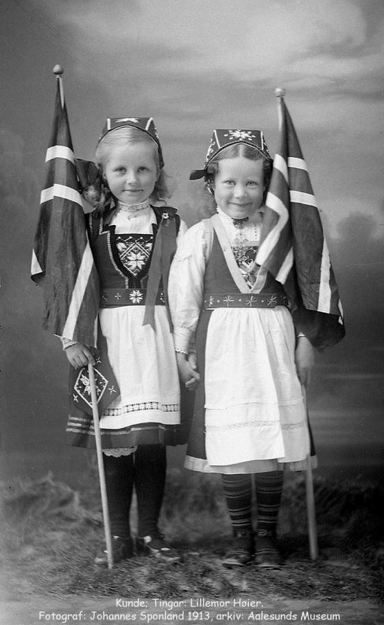 To småjenter hand i hand i fotostudio, smiler mot fotografen i kvar sin bunad. Held i tillegg kvart sitt store norske flagg.