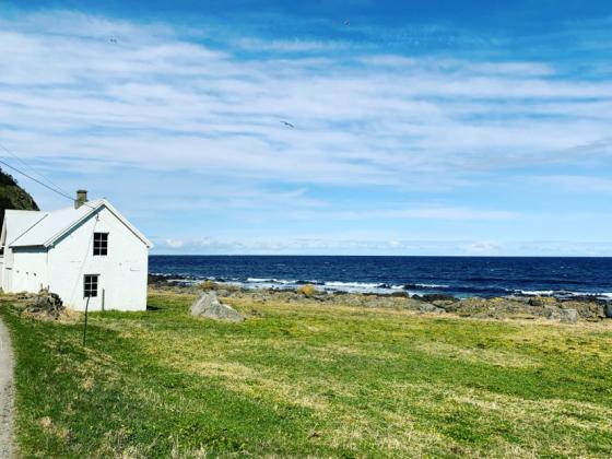 Bilde av ei gammal kvit murbygning på venstre side, i grønt gras, bak i bildet ser ein fjøresteinane og storhavet bak, blå himmel med skyer.