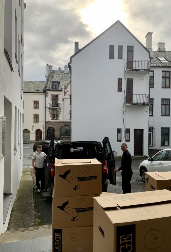 Bilde frå ein bakgård i Ålesund, med høge murhus frå tidleg 1900-tal, overskya ver med litt sol over eit husmøne. Fremst i bildet står det tre stablar med brune flyttekasser, bak dei står det parkert ein liten kassebil, med bakdørene opne, kassene kom frå denne bilen. På sida av bilen, ein på kvar side, står det to menn som smilar til fotografen.