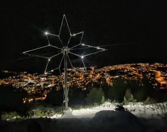 Bilde tatt frå ein liten fjelltopp, ser mot ei stor lysande stjerne som er montert der, med utsikt over ei vinteropplyst bygd
