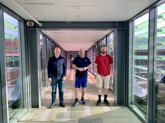 Tre menn står i ein glaskorridor, vende mot fotografen som står i ein ende av korridoren. Alle smiler