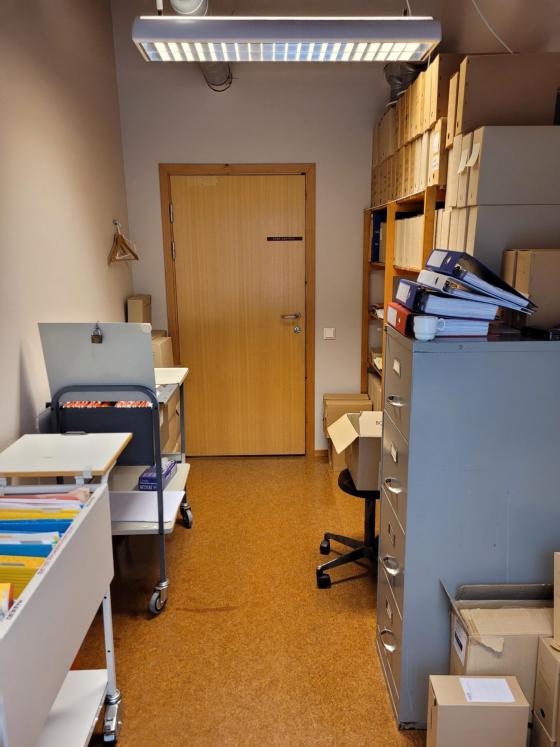 Eit langt smalt rom med arkivskap, traller og trehyller, fylde med arkivboksar, permar og mapper.