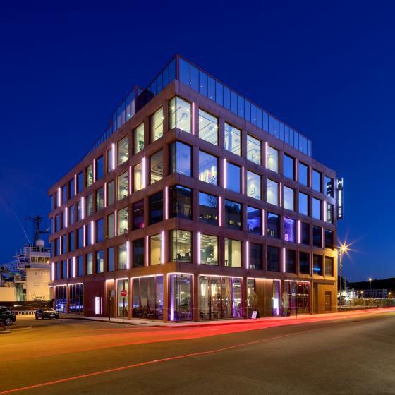 Kveldsbilde i blått lys av eit moderne bygg med mykje vindauge og glasflater. Vegen har ei raud stripe frå ein passerande bil.