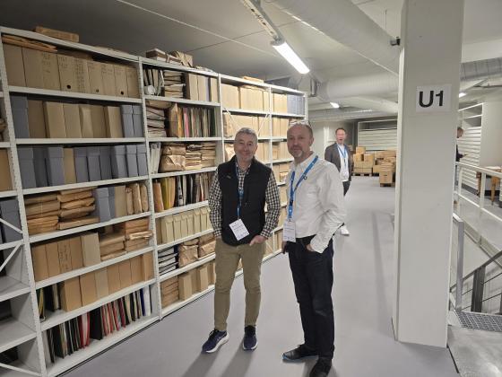 Tre menn står i eit moderne museumsmagasin ved ein fylt reol.