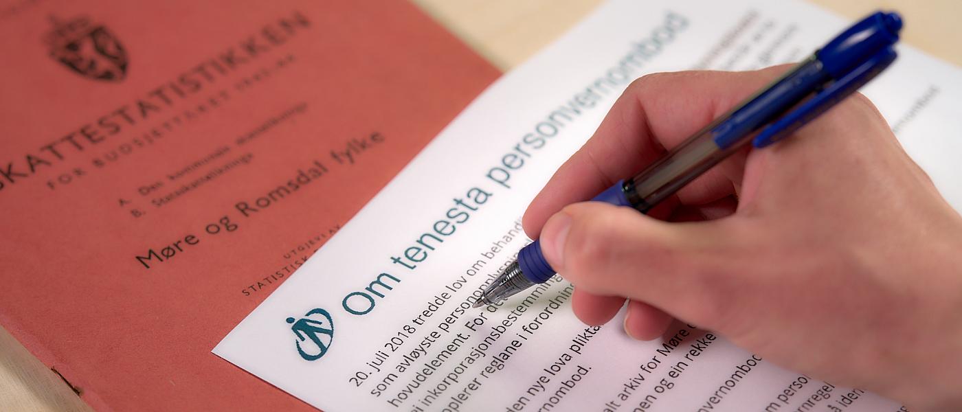 Ei høgrehand som held ein penn ligg oppå eit ark med tekst om tenesta personvernombod - under arket ligg ei raud framside på eit hefte med løvesymbol og teksta skattestatistikk Møre og Romsdal fylke