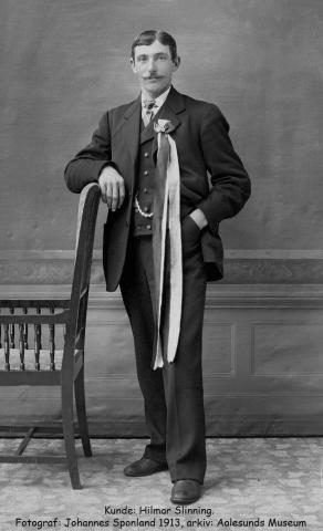 Mann ståande i fotostudio, kviler høgrearm mot toppen av ein stol, har lang 17. maisløyfe og pendress.