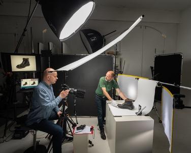 To menn i eit fotostudio med eksterne lyskjelder som lyser på ein stor kloss der det står ein gamal sko. Den eine mannen held i skoen, medan den andre fotograferer den.