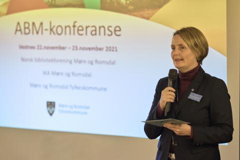 Ei kvinne i dressjakke står med ein mikrofon i handa og ønsker velkomen til konferanse. Bak henne er det ein poiwerpoint-presentasjon der det stpr ABM-konferanse