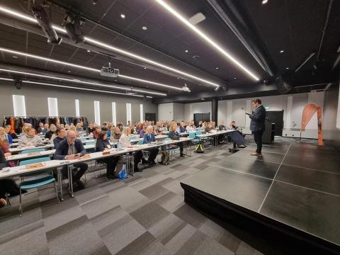 Bilde frå ein konferansesal med golvteppe i koksgrå/svartmelerte ruter. Til høgre i bildet er det eit podum, på podiumet står ein mann og snakkar. I resten av romet er det langbord med konferansedeltakarar.