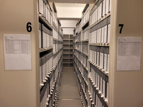 Ein står ute i gangen og ser inn mellom reol 6 og 7, som er fylde med grå arkivboksar, ferdig etikettert.