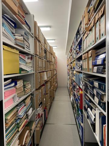 Vi ser inn mellom to arkivreolar, som er fylde med forskjellig arkiv, arkivboksar, permar, bøker og liknande.