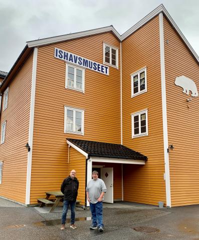 To men står fremst på bildet og ser mot kamera. Bak dei står ein spisebenk inntil eit høgt okerfarga hus med småruta vindauge. På eine veggen er det ei utskjering av ein isbjørn, på den andre står det ishavsmuseet.