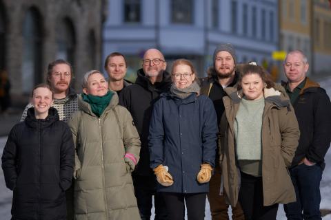 Ei lita gruppe vinterkledde menneske står ute i ei gate med murhus og smiler mot fotografen