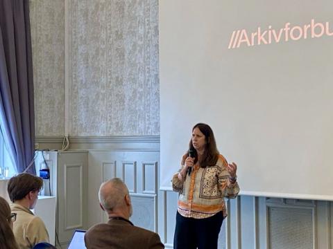 Kvinne med mikrifon som står framfor ei samling sittande menneske