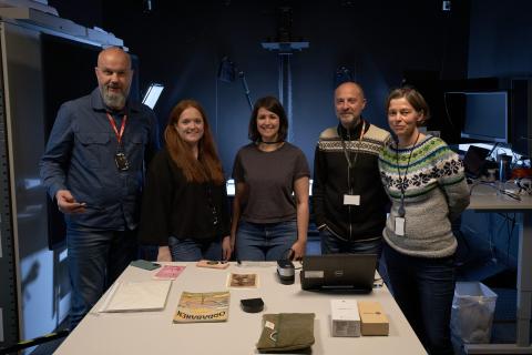 Tre kvinner og to menn i eit black box studio står bak eit kvitt bord med arkivmateriale. Alle smiler mot kamera.