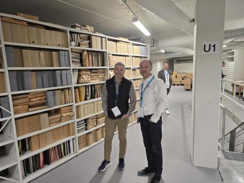 Tre menn står i eit moderne museumsmagasin ved ein fylt reol.