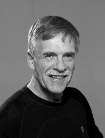 Portrettbilde i svart-kvitt av ein godt voksen korthåra mann i svart trøye, som smiler til fotografen